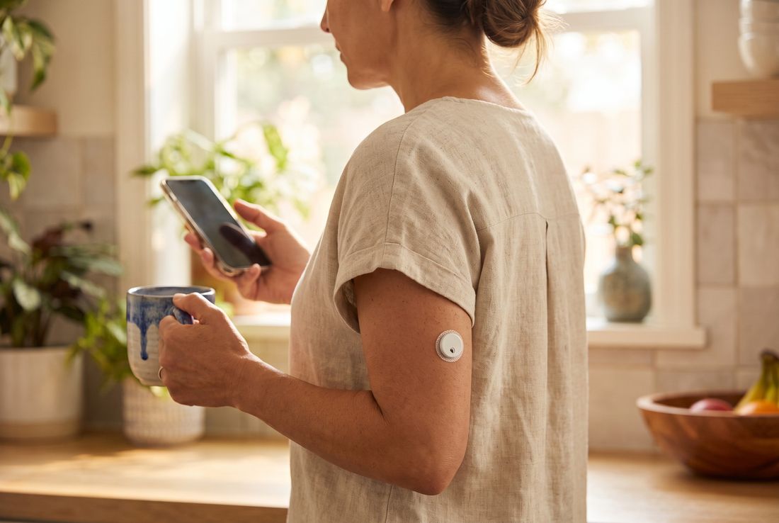 Mujer en su cocina con una taza de café y su celular, con el sensor QiTrax en la parte posterior del brazo